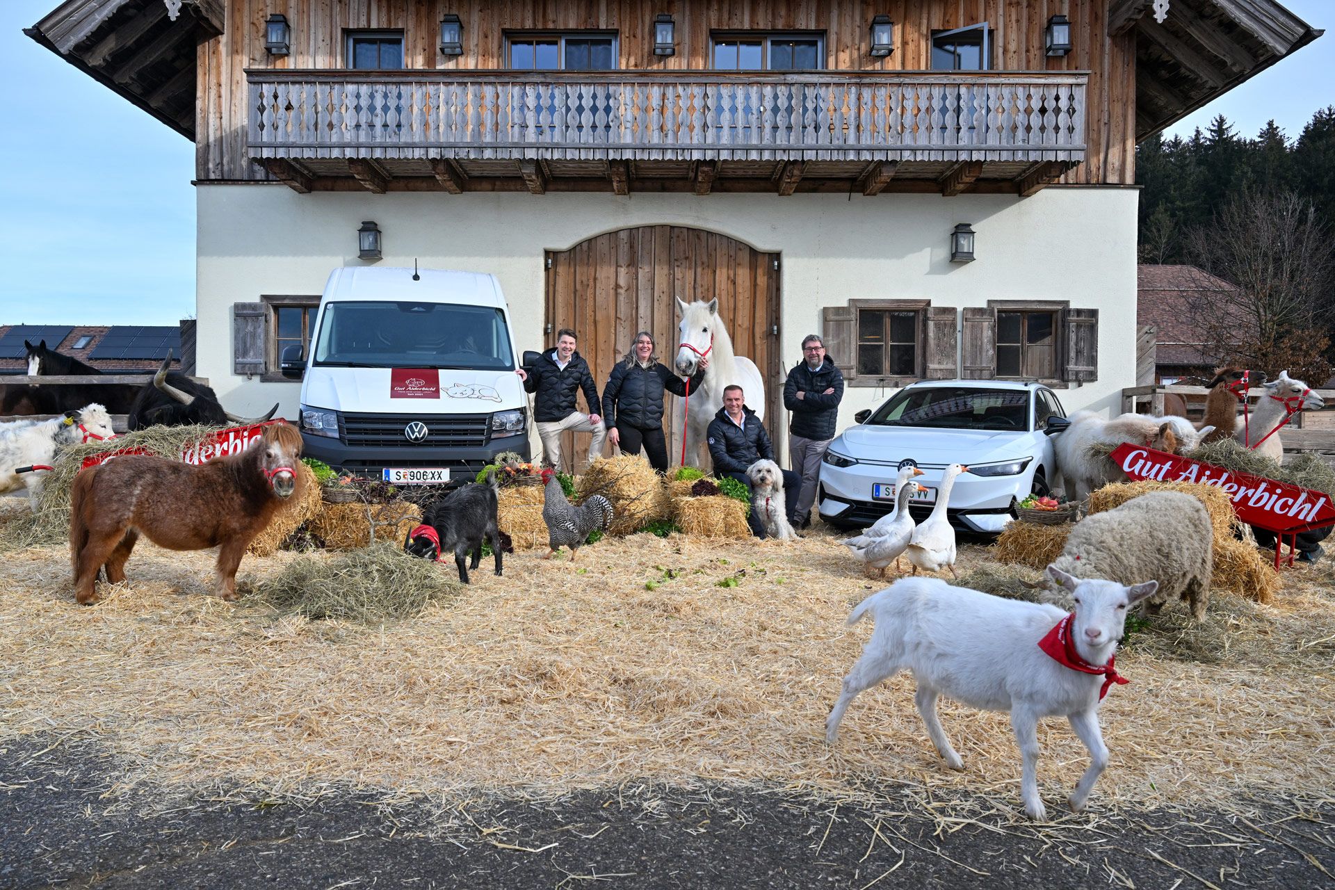 Mehrere Menschen und zahlreiche Tiere, darunter Ziegen, Hühner und Schafe, stehen vor einem alpinen Bauernhaus mit Holzbalkon. Im Hintergrund sind zwei Fahrzeuge geparkt, ein weißer Lieferwagen und ein helles Auto. Einige der Tiere tragen rote Schärpen mit der Aufschrift "Gut Aiderbichl".