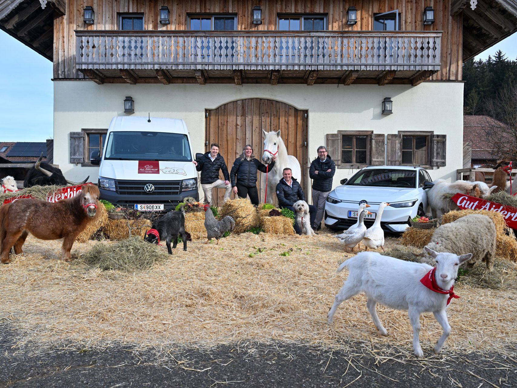 Mehrere Menschen und zahlreiche Tiere, darunter Ziegen, Hühner und Schafe, stehen vor einem alpinen Bauernhaus mit Holzbalkon. Im Hintergrund sind zwei Fahrzeuge geparkt, ein weißer Lieferwagen und ein helles Auto. Einige der Tiere tragen rote Schärpen mit der Aufschrift "Gut Aiderbichl".