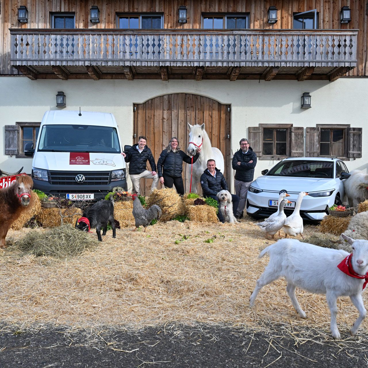 Mehrere Menschen und zahlreiche Tiere, darunter Ziegen, Hühner und Schafe, stehen vor einem alpinen Bauernhaus mit Holzbalkon. Im Hintergrund sind zwei Fahrzeuge geparkt, ein weißer Lieferwagen und ein helles Auto. Einige der Tiere tragen rote Schärpen mit der Aufschrift "Gut Aiderbichl".