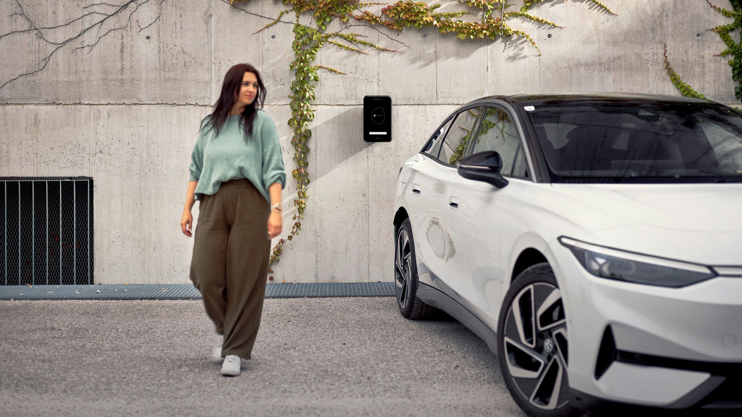 Eine Frau geht in der Nähe eines weißen ID.7, das an einer Wand mit einer Ladestation und Kletterpflanzen geparkt ist. Die Umgebung wirkt urban und modern.