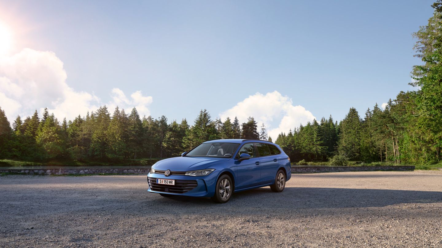VW Passat in blau steht auf einer Kiesfläche, im Hintergrund ein Tannenwald.