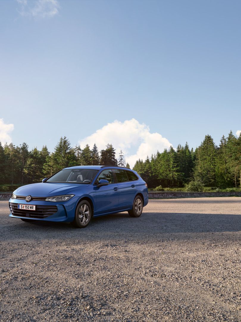 VW Passat in blau steht auf einer Kiesfläche, im Hintergrund ein Tannenwald.