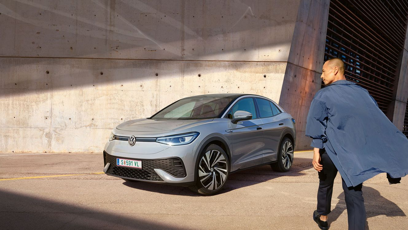 Grauer VW ID.5 parkt vor heller Hauswand, Ansicht schräg rechts von vorn, Mann geht zügig auf das E-Auto zu.