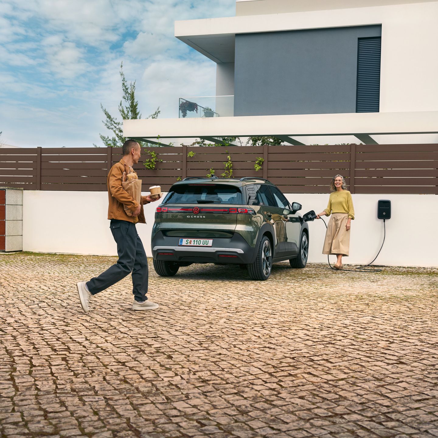 Eine Frau steckt ein Ladekabel einer Wallbox in einen grünen VW ID. Cross, ein Mann geht auf sie zu und hat Kaffeebecher in der Hand und Brot unter dem Arm.