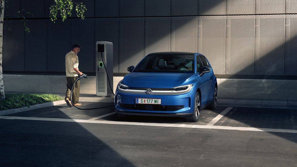 Ein blauer ID. Polo steht auf einem Parkplatz neben einer Ladesäule. Im Hintergrund sind ein Gebäude und ein Baum zu sehen.