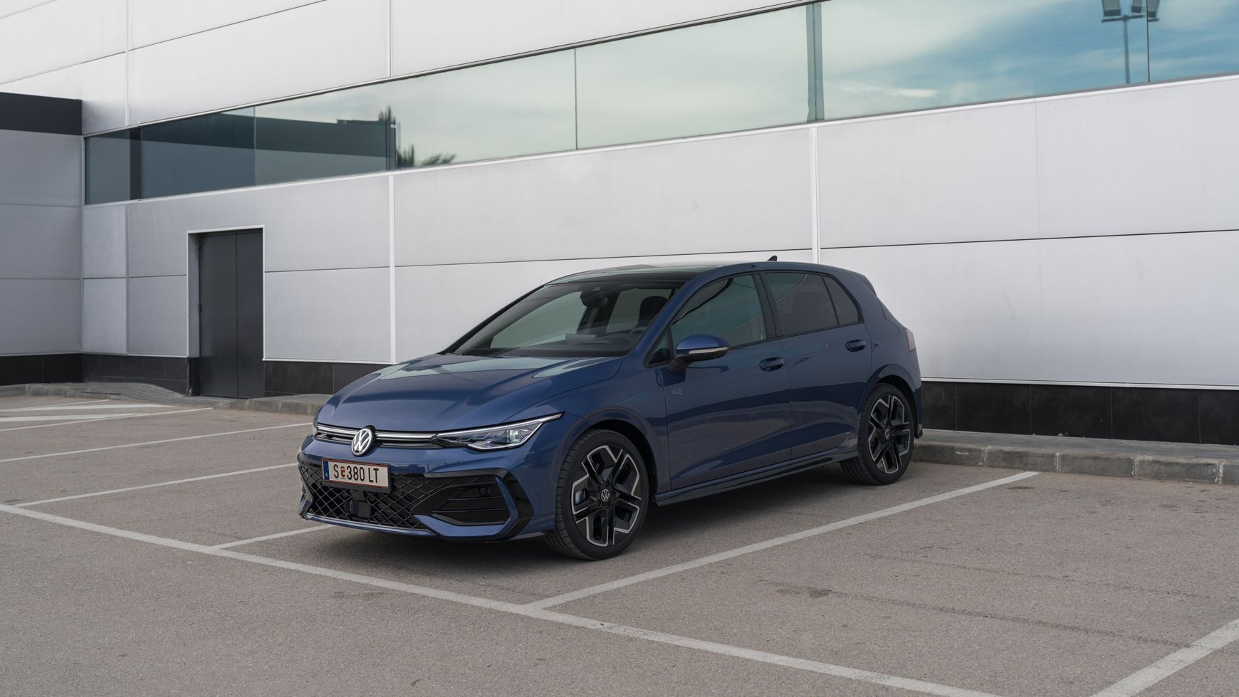 Ein blauer VW Golf ist auf einem leeren Parkplatz vor einem modernen, grauen Gebäude mit großen Fenstern geparkt. Es herrscht bewölktes Wetter. Der Wagen steht diagonal zur Kamera.