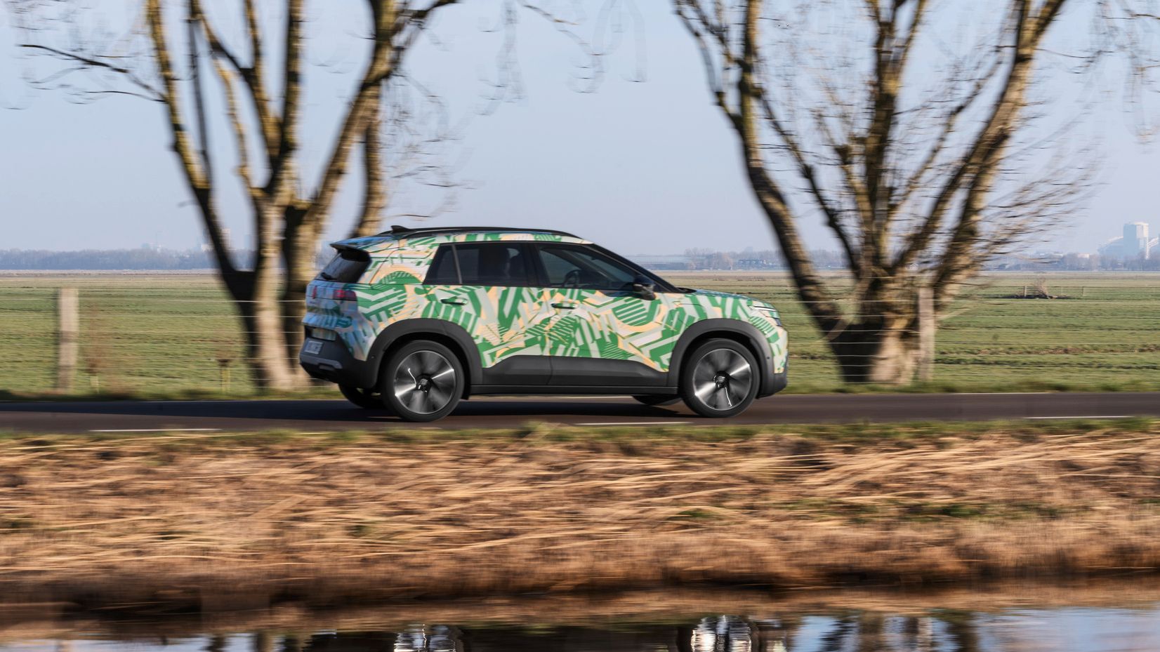 Ein SUV fährt auf einer ländlichen Straße vorbei an kahlen Bäumen und einem Wassergraben. Das Auto ist mit einem bunten, tropischen Muster bemalt. Im Hintergrund ist eine grüne Wiese und wolkenloser Himmel zu sehen.