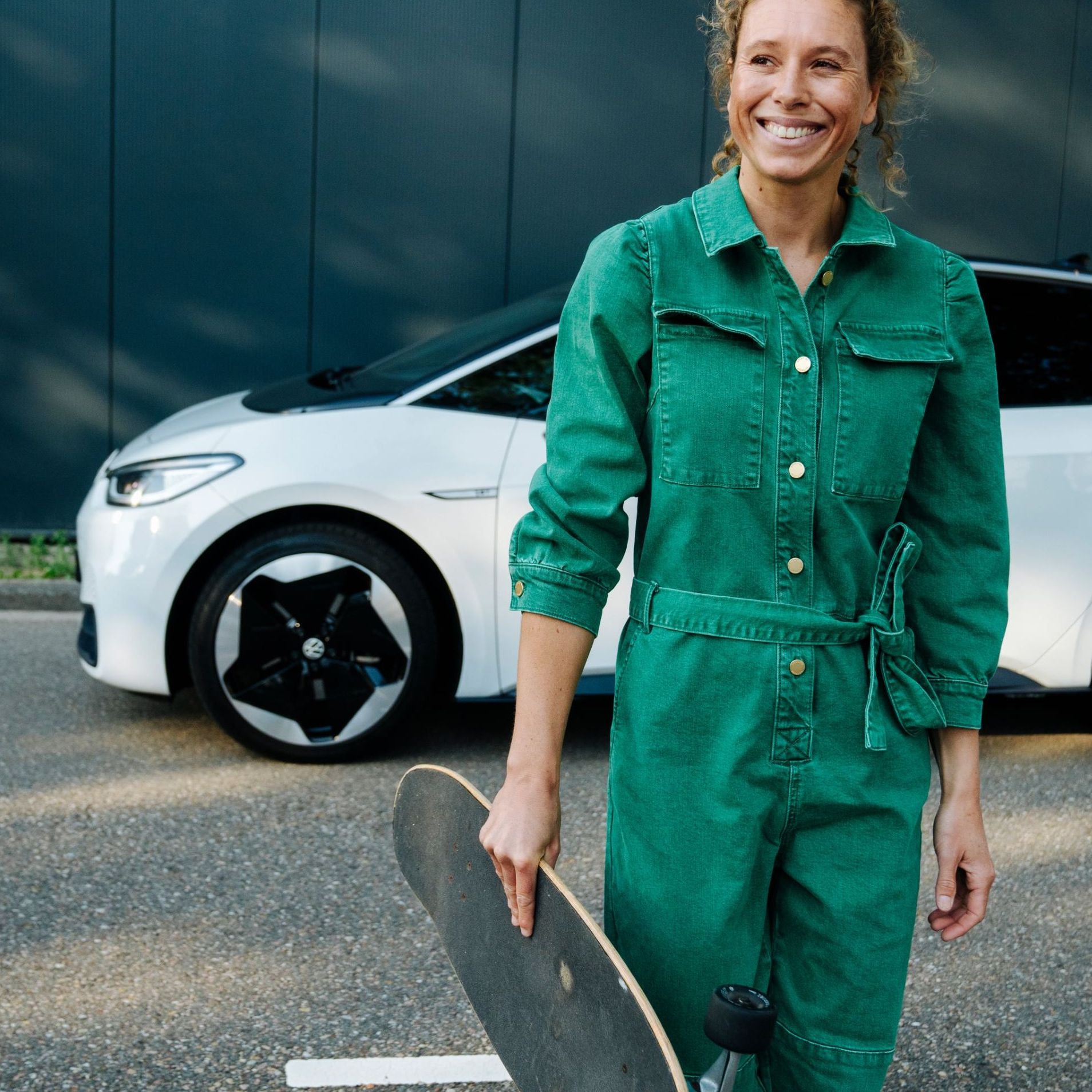 Eine Person in einem grünen Overall hält ein Skateboard und steht auf einer Straße vor einem weißen Auto.