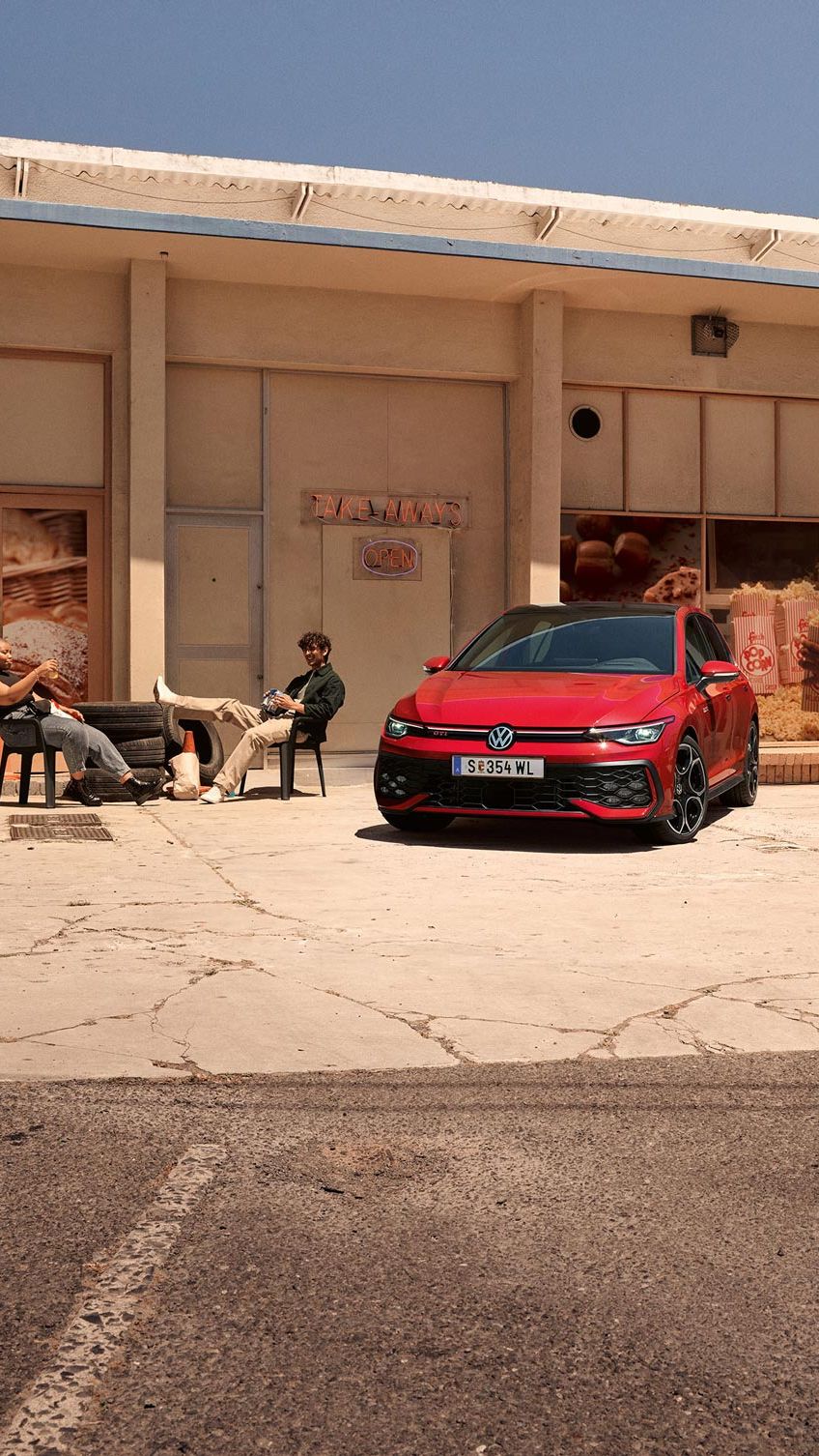 Zwei junge Männer sitzen vor einem Geschäft neben einem roten VW Golf GTI.