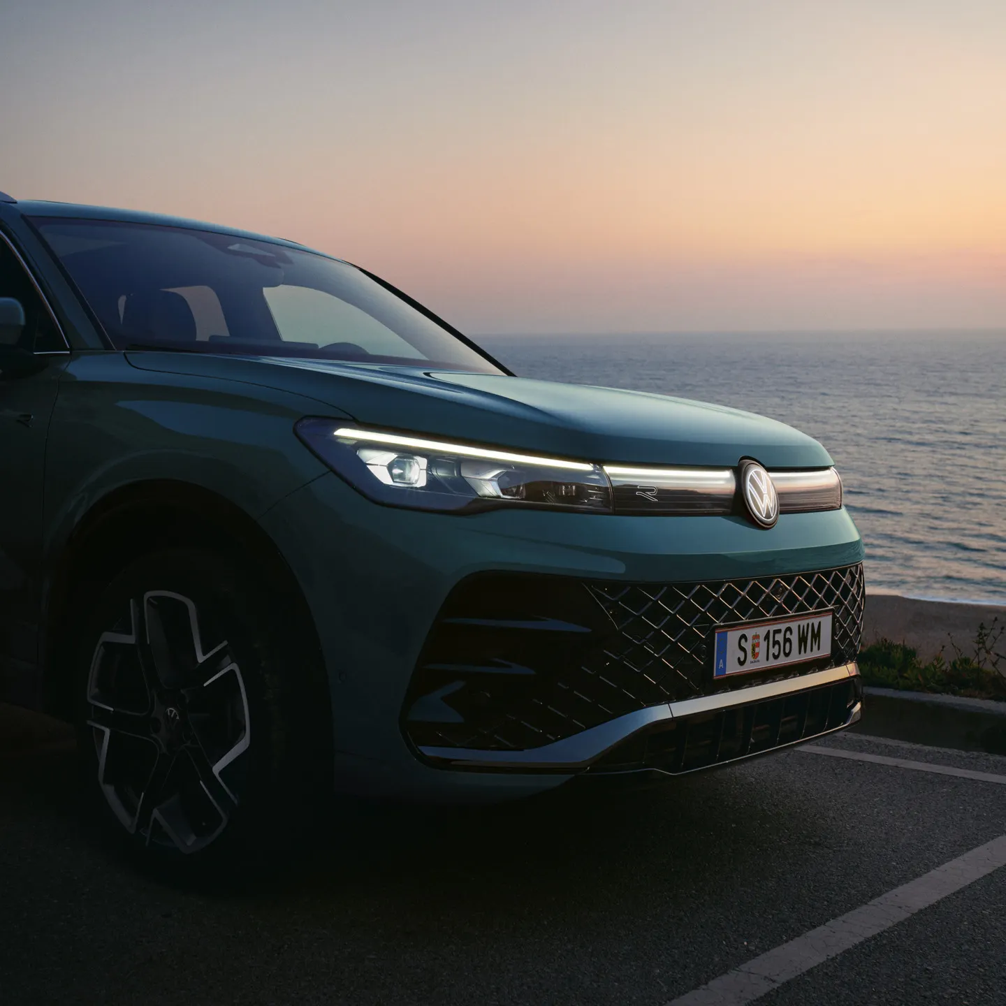 Front-Lichtsignatur eines blauen VW Tiguan am Meer beim Sonnenuntergang.