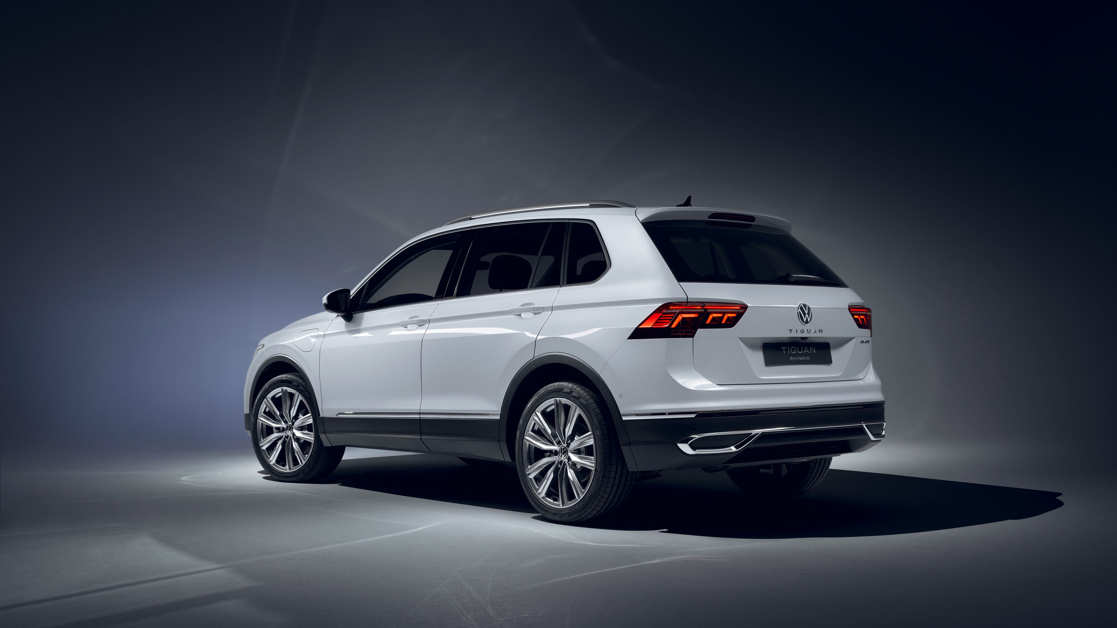 Ein silberner Volkswagen Tiguan SUV steht in einem dunklen Raum. Das Auto ist seitlich und von hinten zu sehen. Die Beleuchtung hebt das elegante Design und die aerodynamischen Linien hervor.