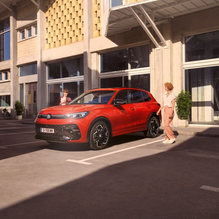Eine junge Frau und ein junger Mann gehen zu ihrem roten Tiguan R-Line, der vor einem Bürogebäude auf einem Parkplatz steht.