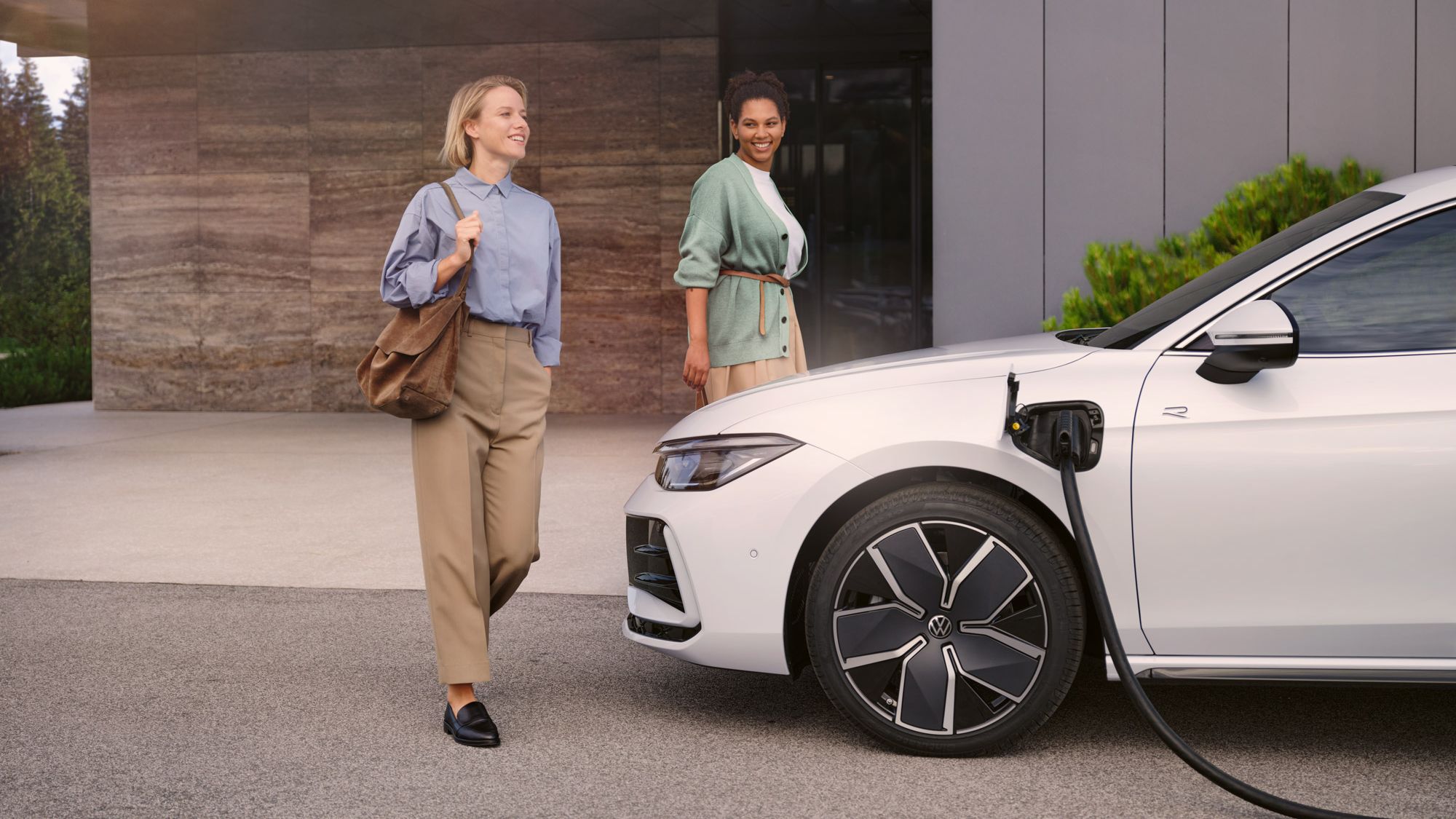 Weißer VW Passat steht mit eingestecktem Ladekabel vor einem Haus, zwei Frauen gehen auf den Wagen zu.