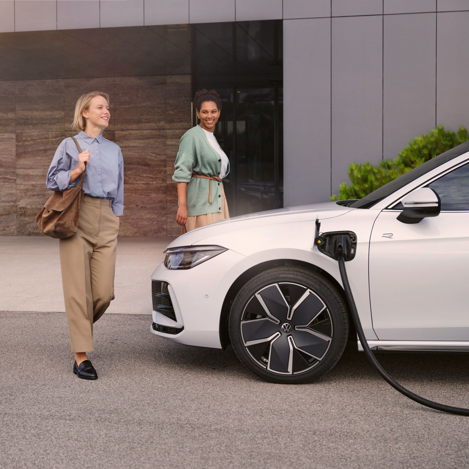 Weißer VW Passat steht mit eingestecktem Ladekabel vor einem Haus, zwei Frauen gehen auf den Wagen zu.