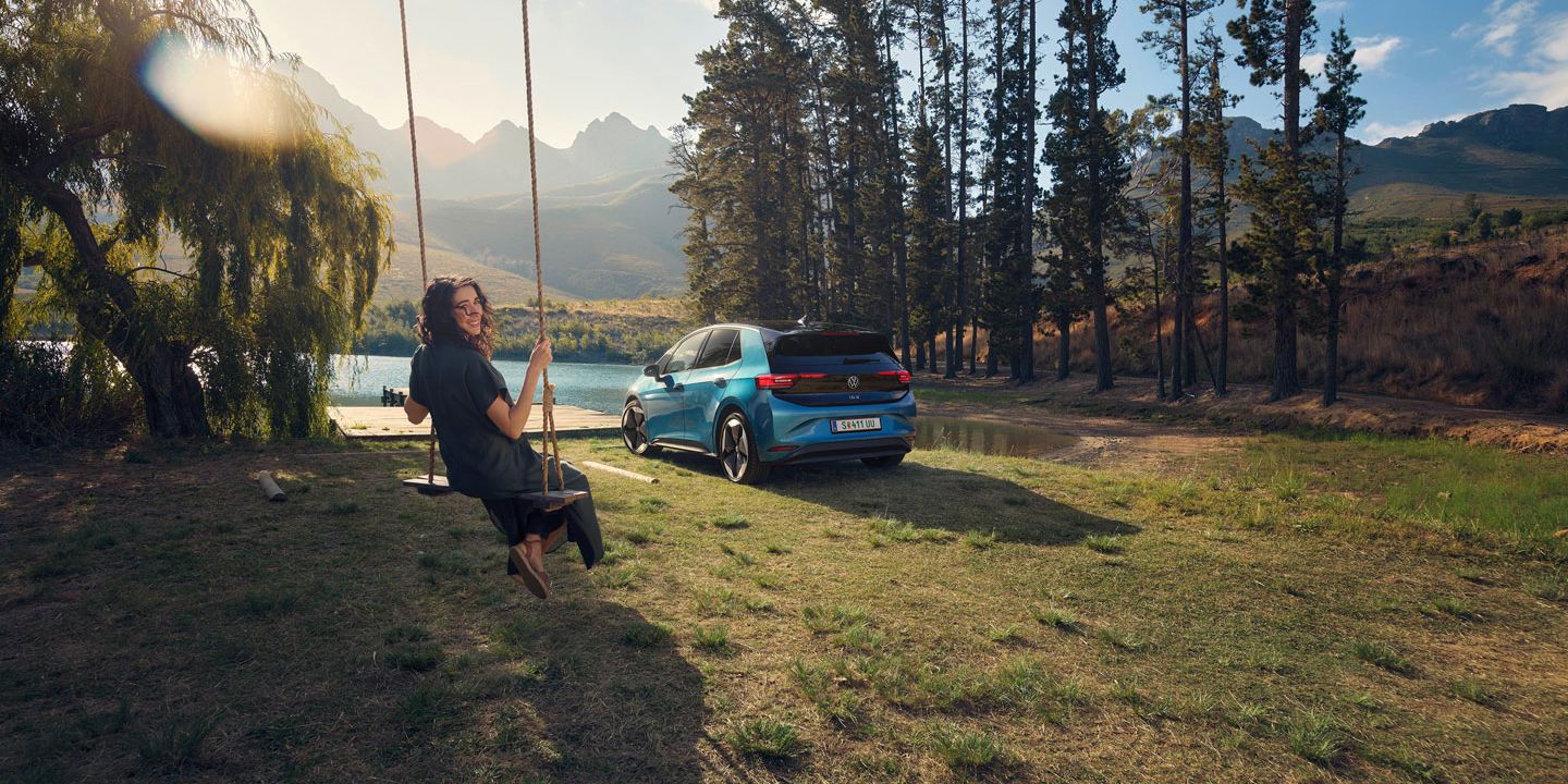 Person schaukelt auf einer Schaukel in einer landschaftlich reizvollen Gegend mit Bergen und einem See im Hintergrund. Neben der Person steht ein blaues Auto vor einer Reihe von Bäumen. Die Szene vermittelt ein Gefühl von Freizeit und Natur.