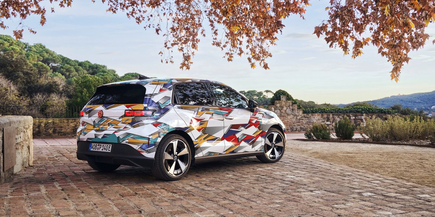 Ein ID. Polo mit bunter geometrischer Folierung steht auf einem gepflasterten Hof. Im Hintergrund sind Hügel mit Bäumen sichtbar, und der Himmel ist teilweise bewölkt. Ein Baum mit Herbstblättern ragt von oben in das Bild hinein.