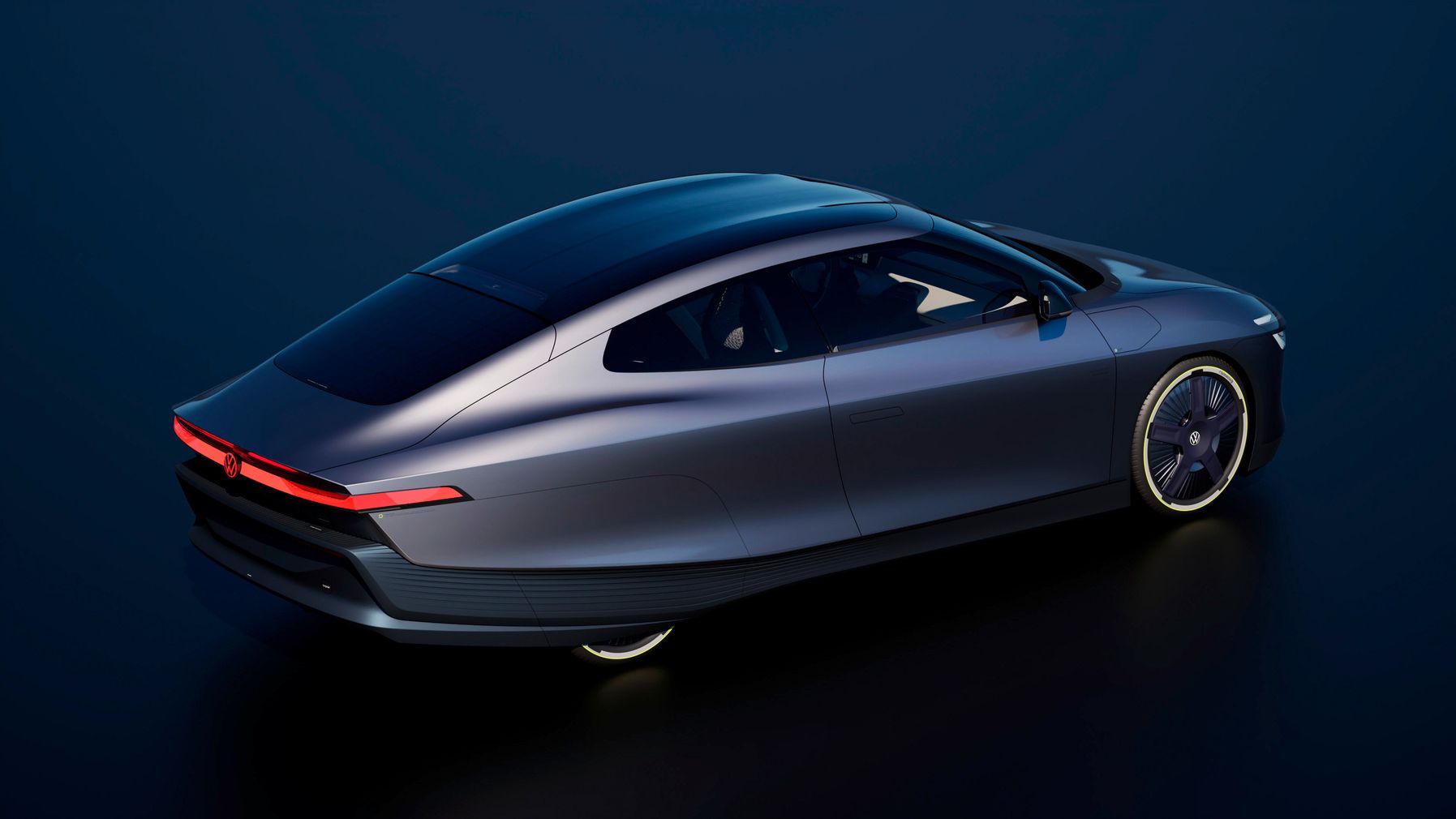 Schlankes, dunkelgraues Elektroauto mit futuristischem, tropfenförmigem Design, fotografiert aus schräger Vogelperspektive auf dunklem Hintergrund. Die Karosserie wirkt sehr aerodynamisch mit fließenden Linien, einem durchgehenden roten LED-Lichtband am Heck und weit nach hinten gezogener Glasdachfläche. Die Felgen sind groß und modern gestaltet, teilweise dezent beleuchtet.