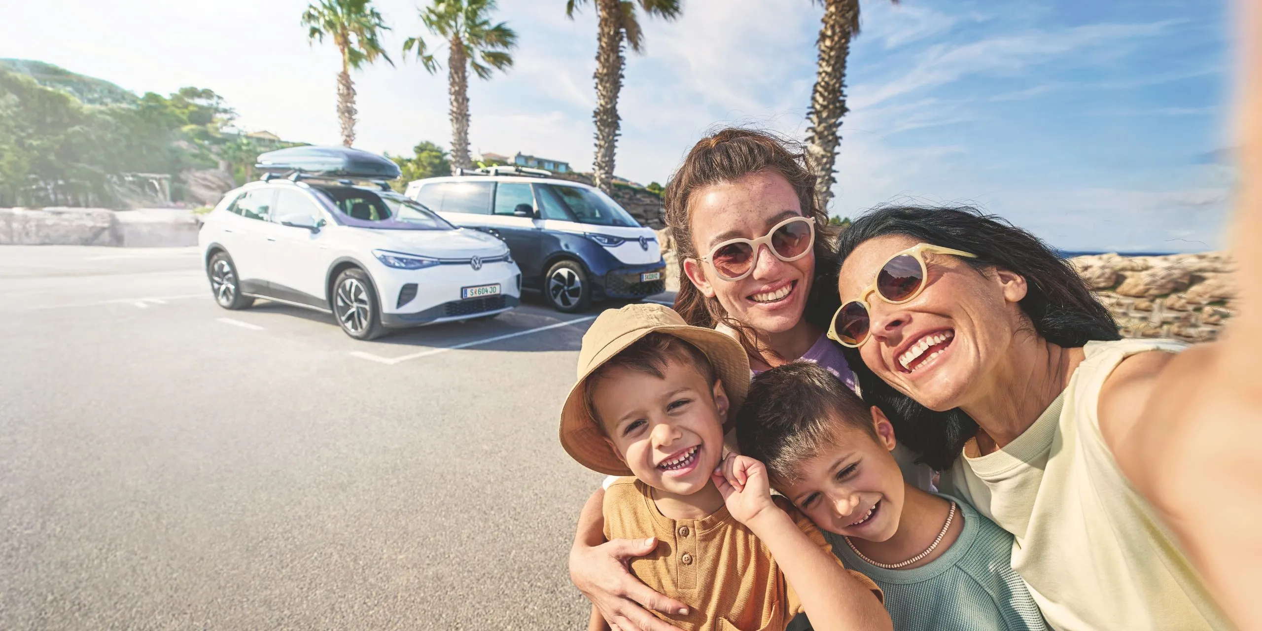 Eine Familie macht ein Selfie auf einem sonnigen Parkplatz mit Palmen im Hintergrund. Im Hintergrund stehen zwei geparkte Volkswagen mit Gepäck auf dem Dach.