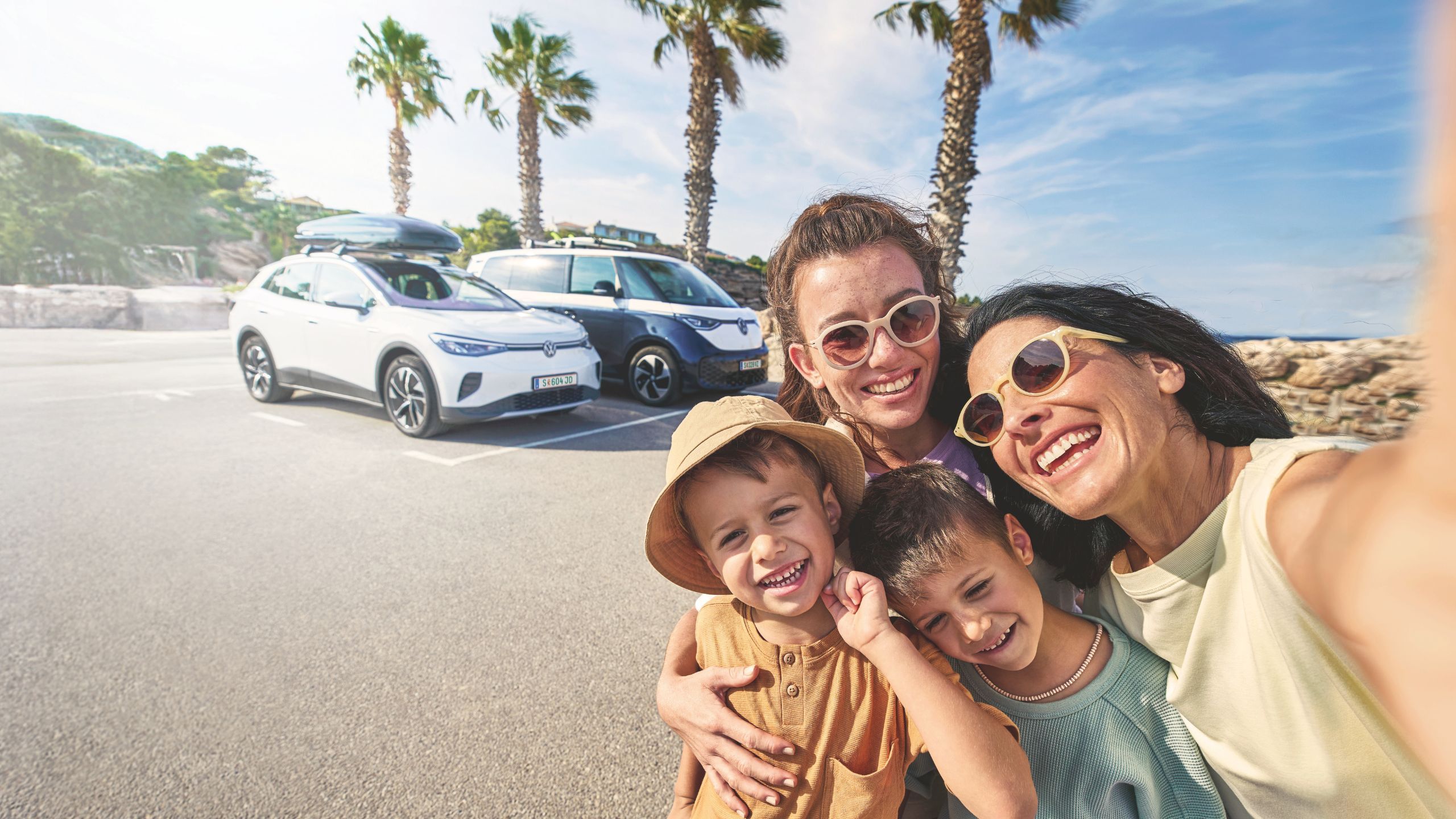 Eine Familie macht ein Selfie auf einem sonnigen Parkplatz mit Palmen im Hintergrund. Im Hintergrund stehen zwei geparkte Volkswagen mit Gepäck auf dem Dach.