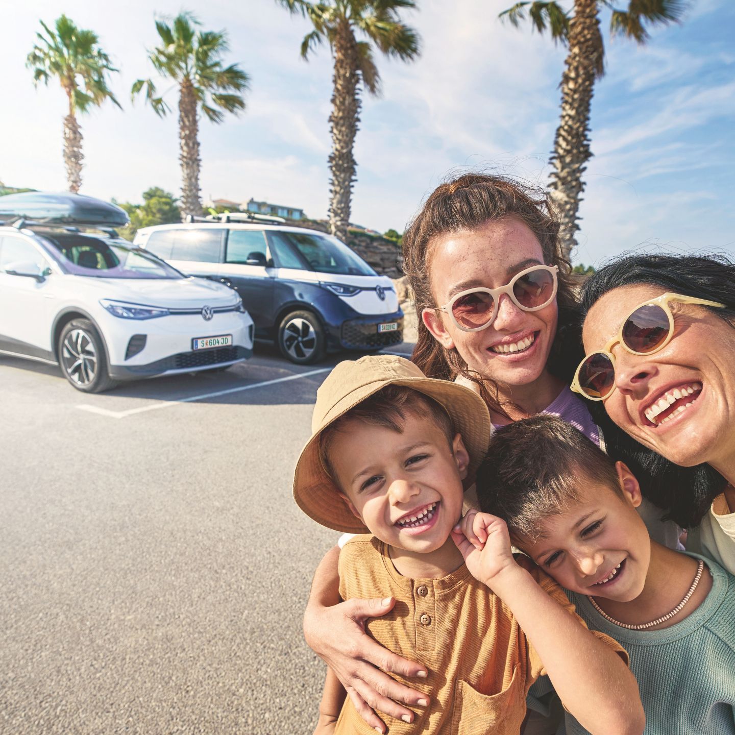 Eine Familie macht ein Selfie auf einem sonnigen Parkplatz mit Palmen im Hintergrund. Im Hintergrund stehen zwei geparkte Volkswagen mit Gepäck auf dem Dach.