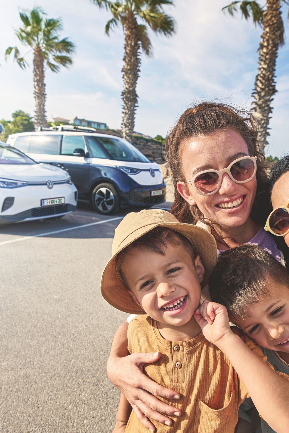 Eine Familie macht ein Selfie auf einem sonnigen Parkplatz mit Palmen im Hintergrund. Im Hintergrund stehen zwei geparkte Volkswagen mit Gepäck auf dem Dach.