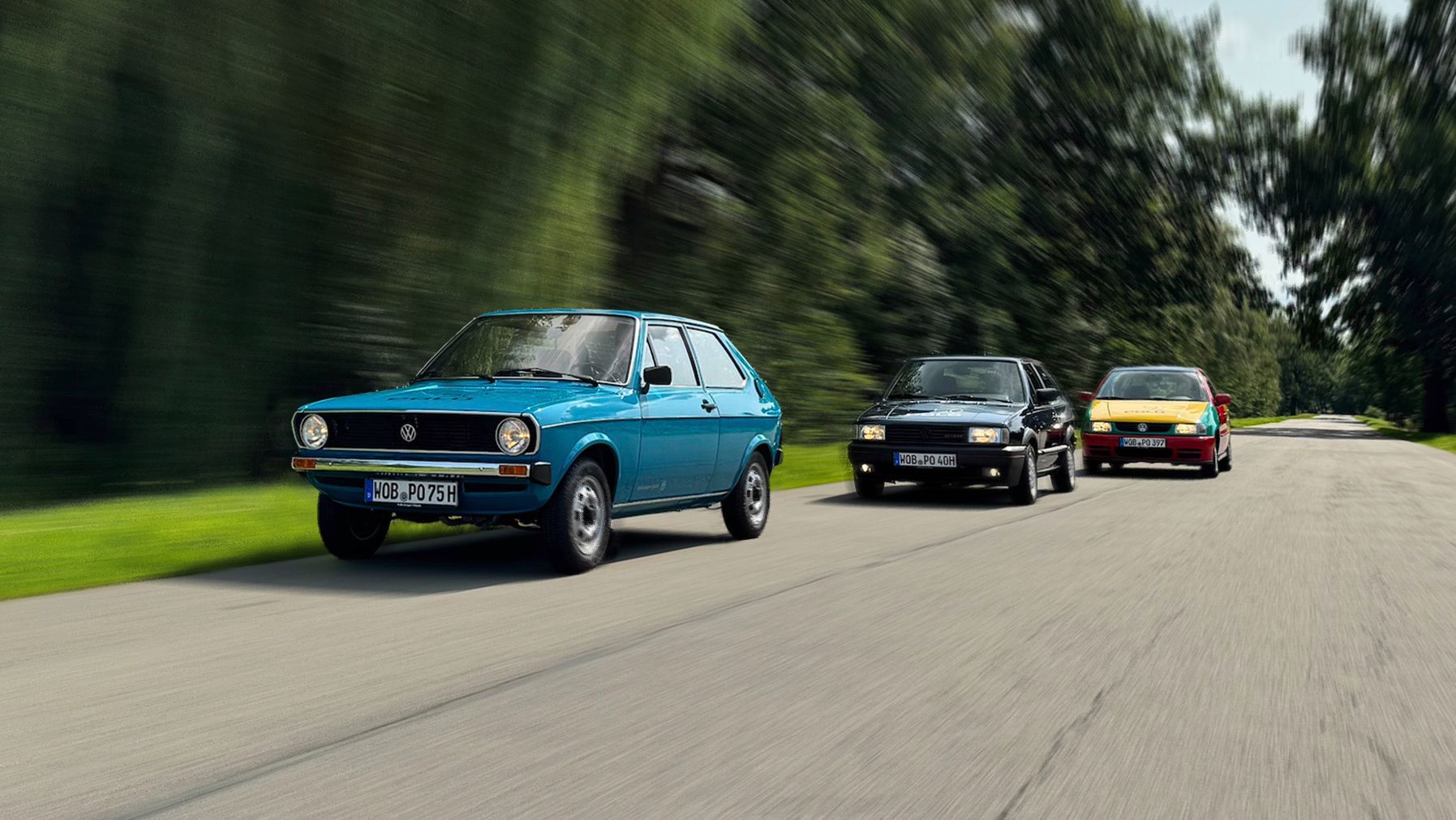 Drei VW Polos fahren hintereinander auf einer Landstraße durch eine grüne, waldige Landschaft. Das vordere Auto ist ein blauer Polo Mk1, das in der Mitte ein schwarzer Polo Mk2 und das hintere ein gelb-rot-grüner Polo Harlekin.