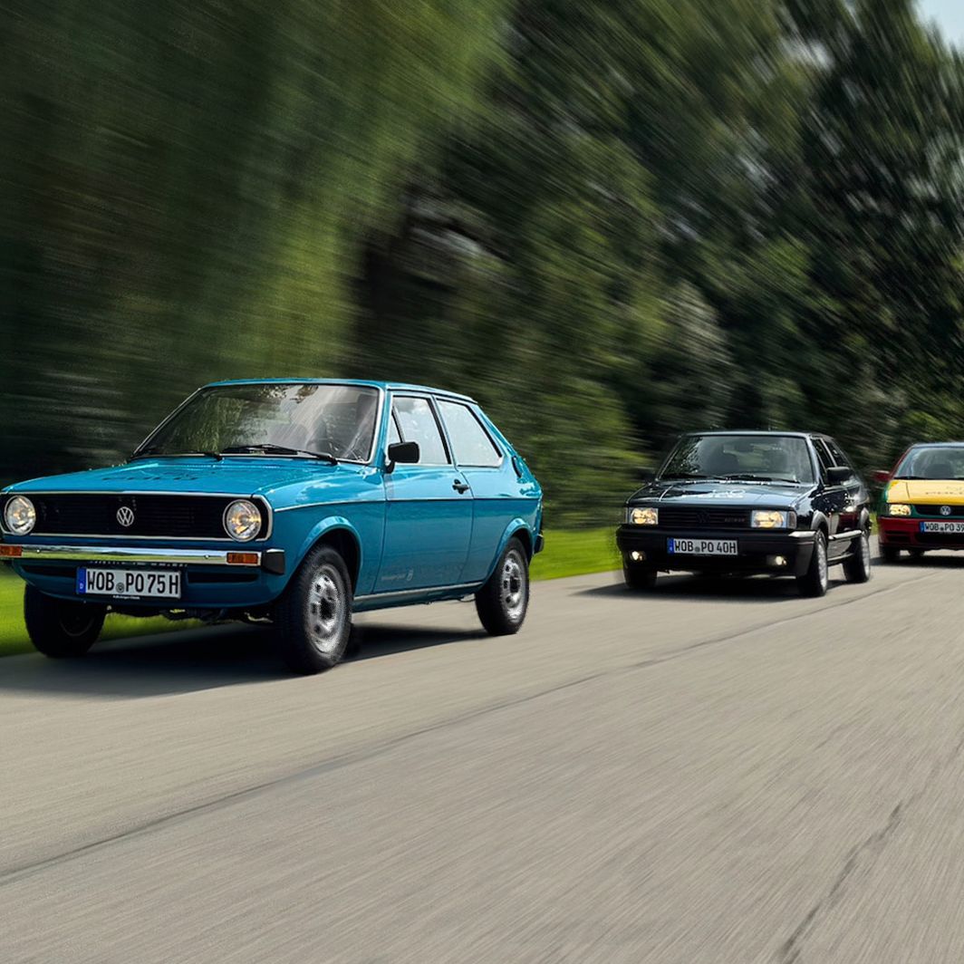 Drei VW Polos fahren hintereinander auf einer Landstraße durch eine grüne, waldige Landschaft. Das vordere Auto ist ein blauer Polo Mk1, das in der Mitte ein schwarzer Polo Mk2 und das hintere ein gelb-rot-grüner Polo Harlekin.