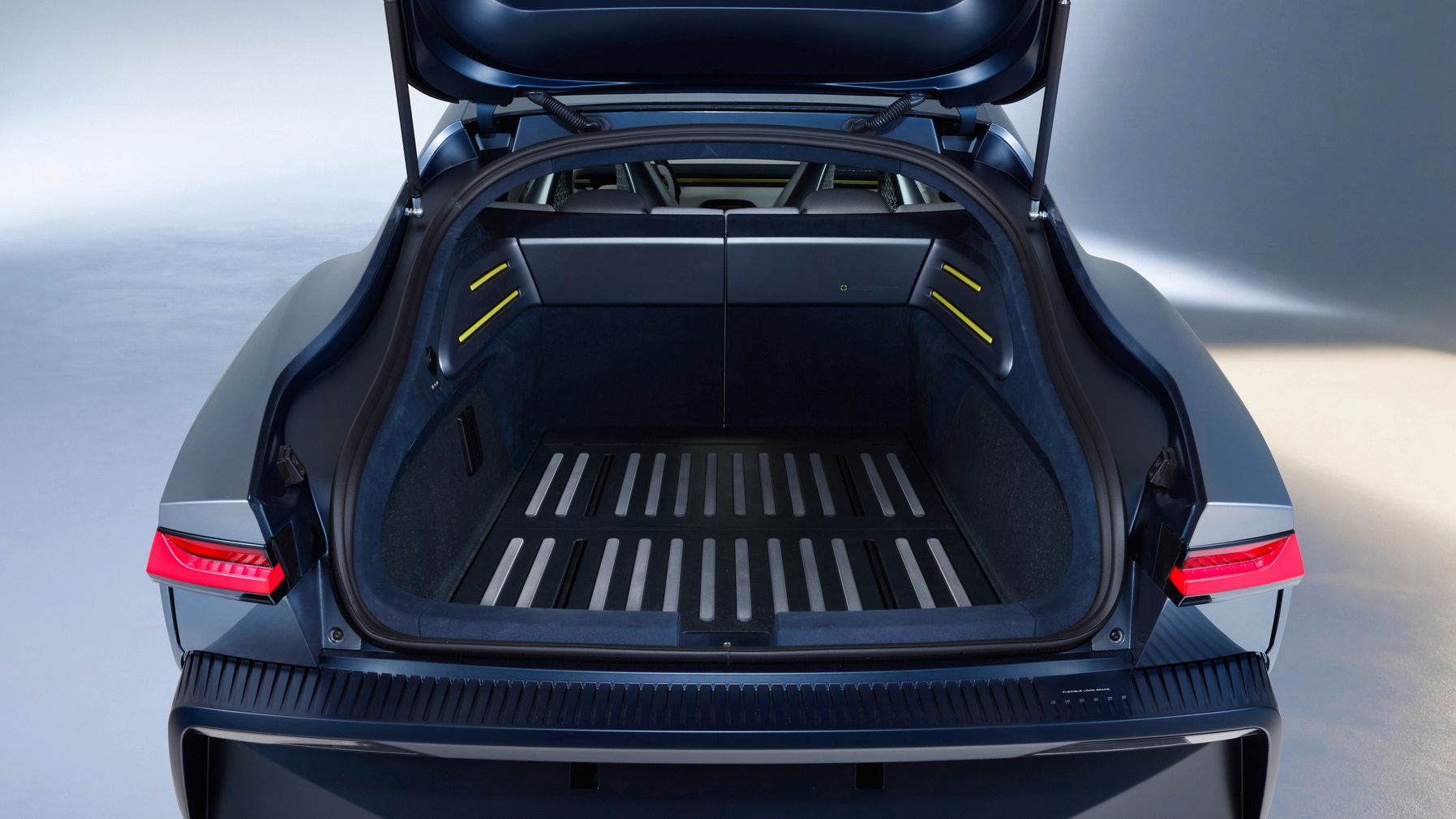 Offener Kofferraum eines modernen Elektroautos mit großem, flachem Ladeabteil und beleuchteten Seitenwänden. Die Rückleuchten sind schmal und futuristisch gestaltet, das Interieur wirkt minimalistisch und hochwertig. Im Boden des Kofferraums sind mehrere helle Streifen eingelassen, die Struktur oder Befestigungsschienen andeuten.