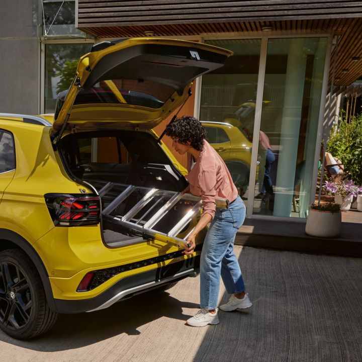 Eine junge Frau schiebt eine Leiter in den Kofferraum eines gelben VW T-Cross.