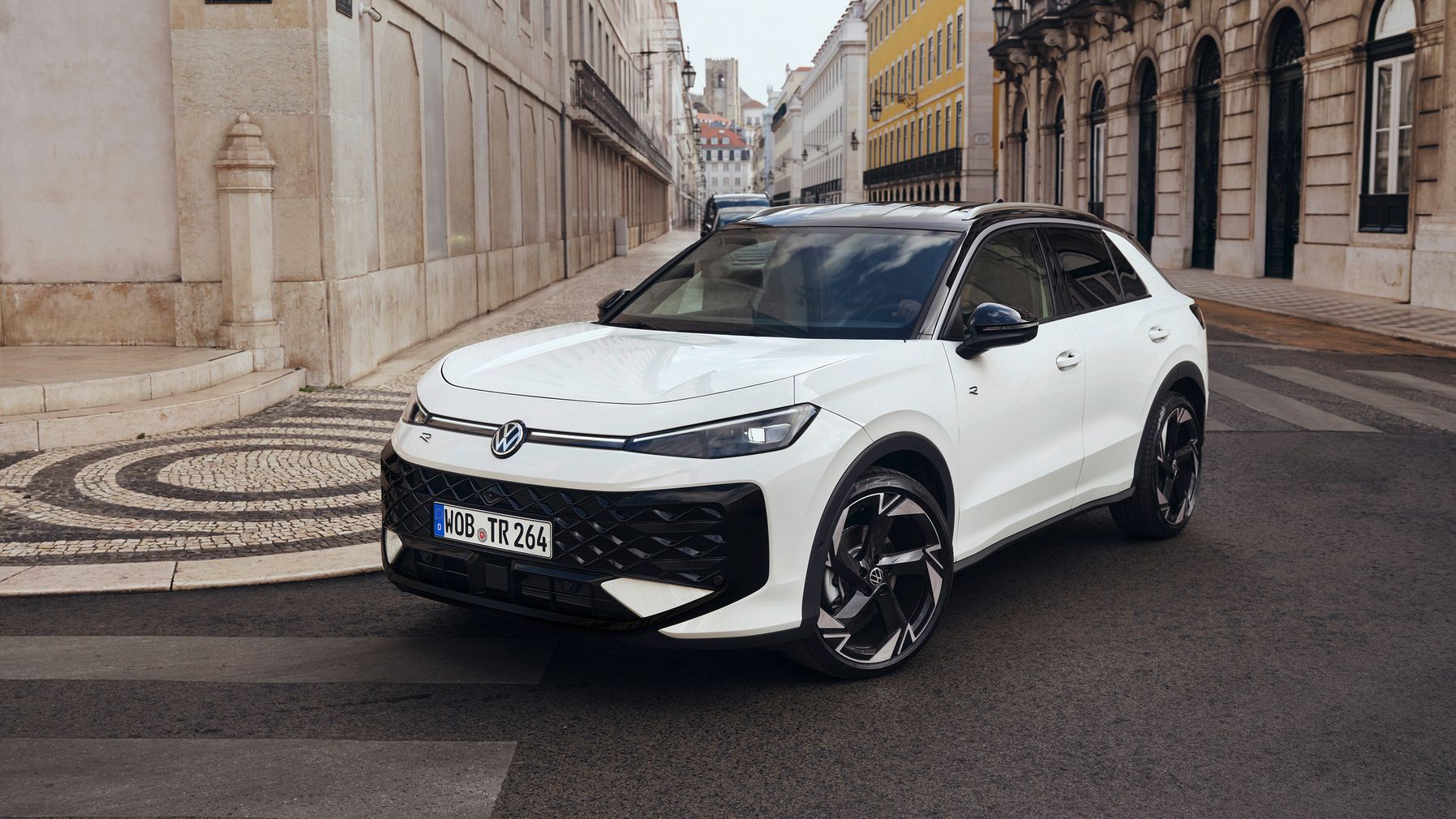 Ein weißer Volkswagen steht auf einer gepflasterten Straße in einer städtischen Umgebung. Im Hintergrund sind Gebäude im europäischen Architekturstil zu sehen.