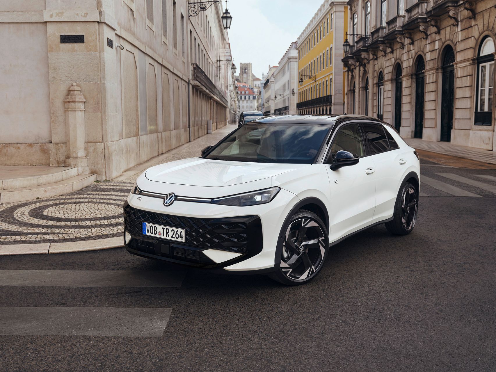 Ein weißer Volkswagen steht auf einer gepflasterten Straße in einer städtischen Umgebung. Im Hintergrund sind Gebäude im europäischen Architekturstil zu sehen.