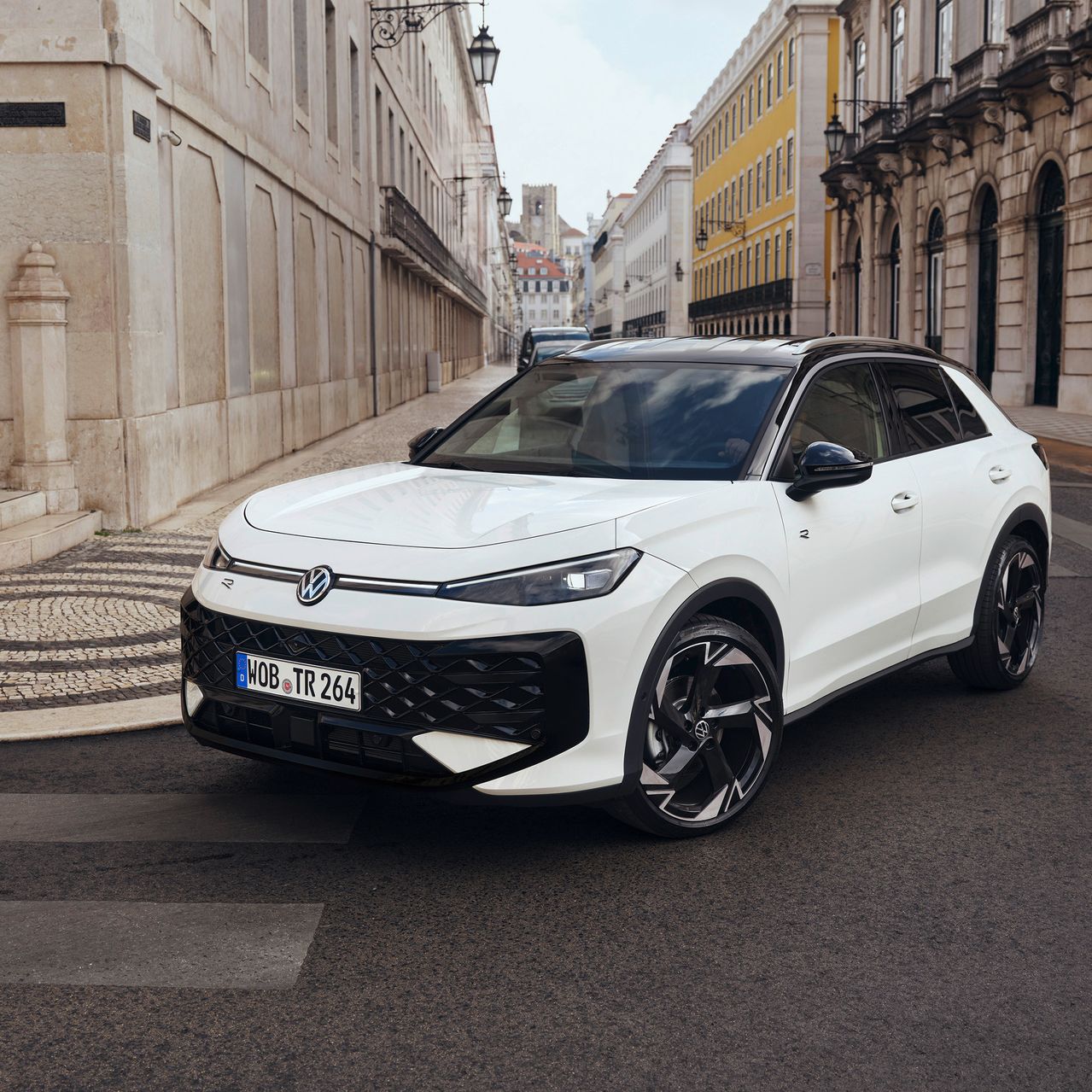 Ein weißer Volkswagen steht auf einer gepflasterten Straße in einer städtischen Umgebung. Im Hintergrund sind Gebäude im europäischen Architekturstil zu sehen.