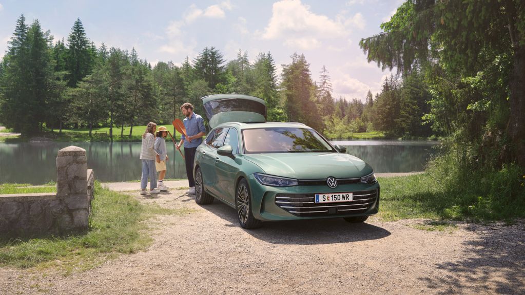 Der neue VW Passat Elegance in Maripositgrün Metallic parkt an einem Badesee mit geöffnetem Kofferraum. Ein Mann verteilt Freitzeitspielzeug an zwei Kinder.