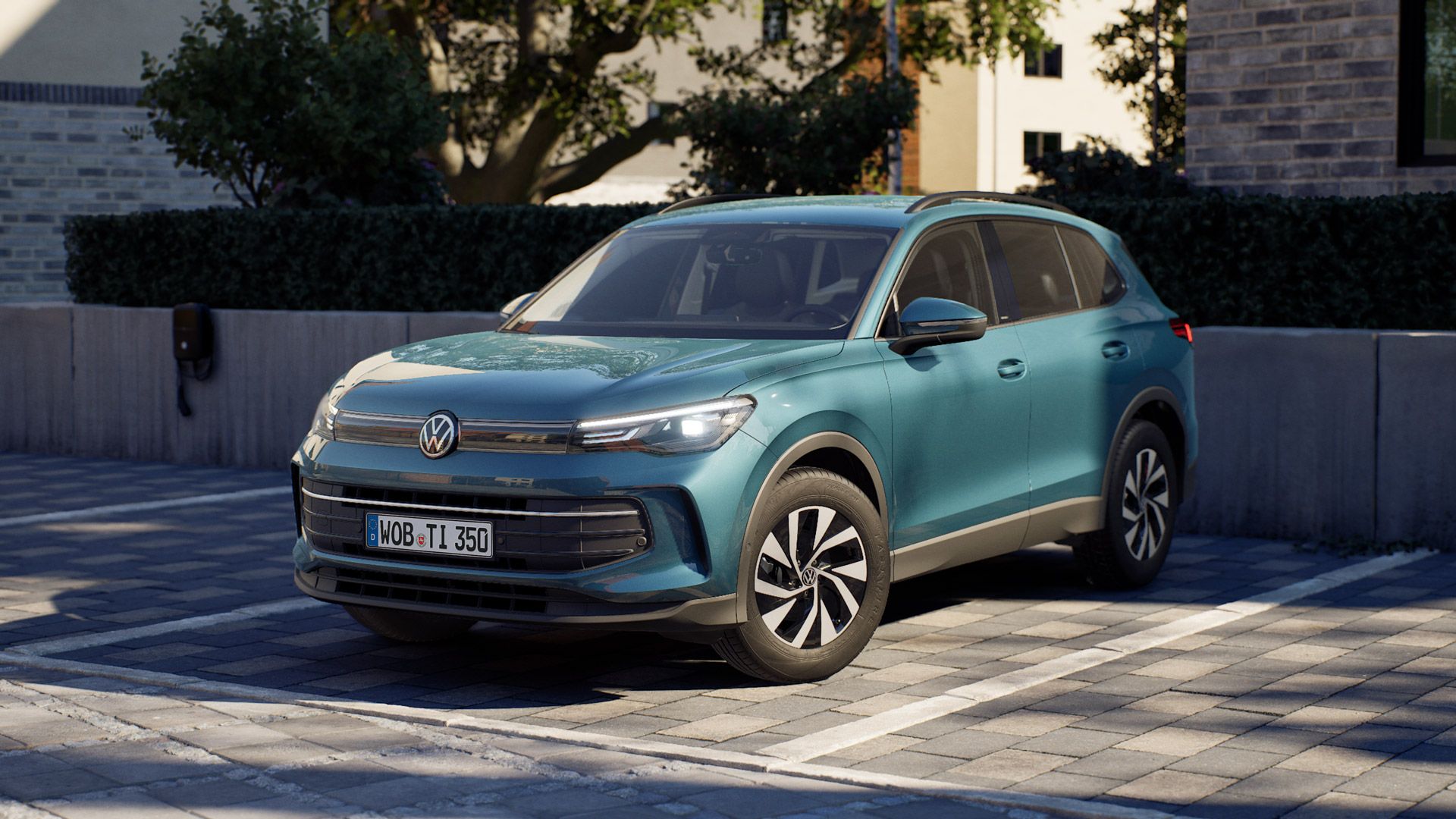 Ein blauer VW Tiguan steht auf einem gepflasterten Parkplatz in einem Wohngebiet, umgeben von modernen Gebäuden und Bäumen. Das Auto ist leicht schräg von vorne fotografiert, sodass Front und Fahrerseite gut sichtbar sind. Die Szene wirkt sonnig und ruhig.