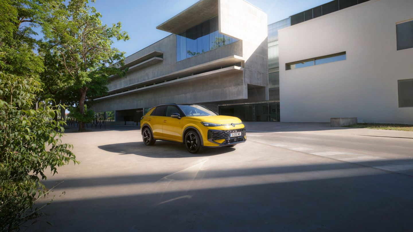 Ein gelber T-Roc R steht vor einer modernen, geometrisch gestalteten Betonarchitektur. Das Gebäude hat breite Fenster und schwebende Dächer. Im Hintergrund sind grüne Bäume zu sehen.