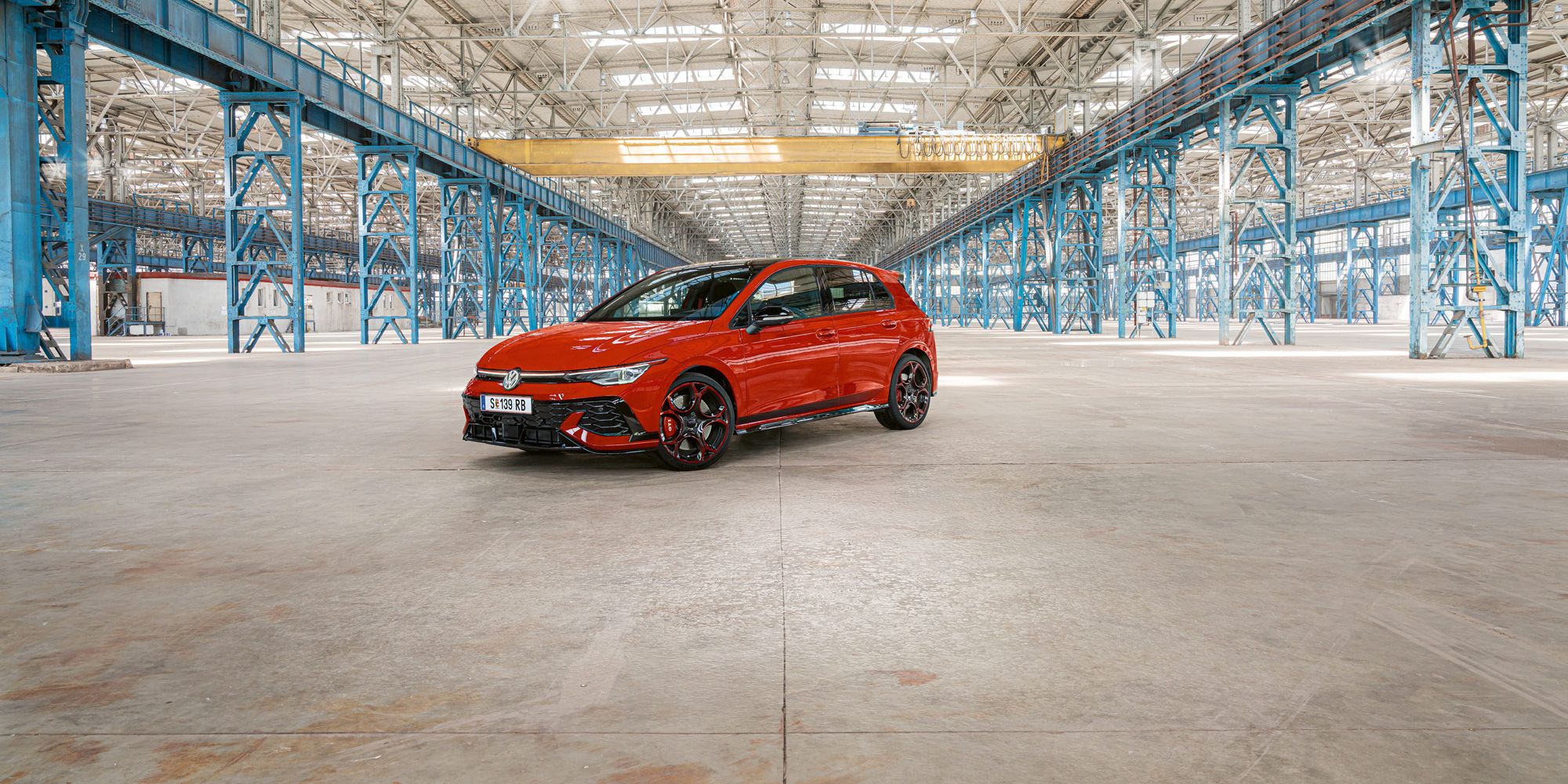 Ein roter Golf GTI steht in einer großen, leeren Industriehalle mit blauen Stahlträgern. Das Dach der Halle ist aus Glas und lässt viel Licht herein.