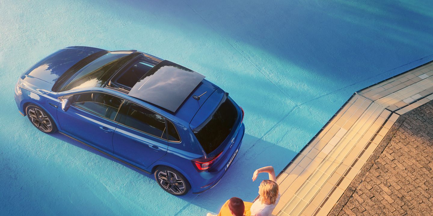  Blick von oben auf blauen VW Polo im Sonnenschein mit geöffnetem Panorama-Ausstell- /Schiebedach in einem leeren Pool. Zwei Personen sitzen am Rand des Pools.