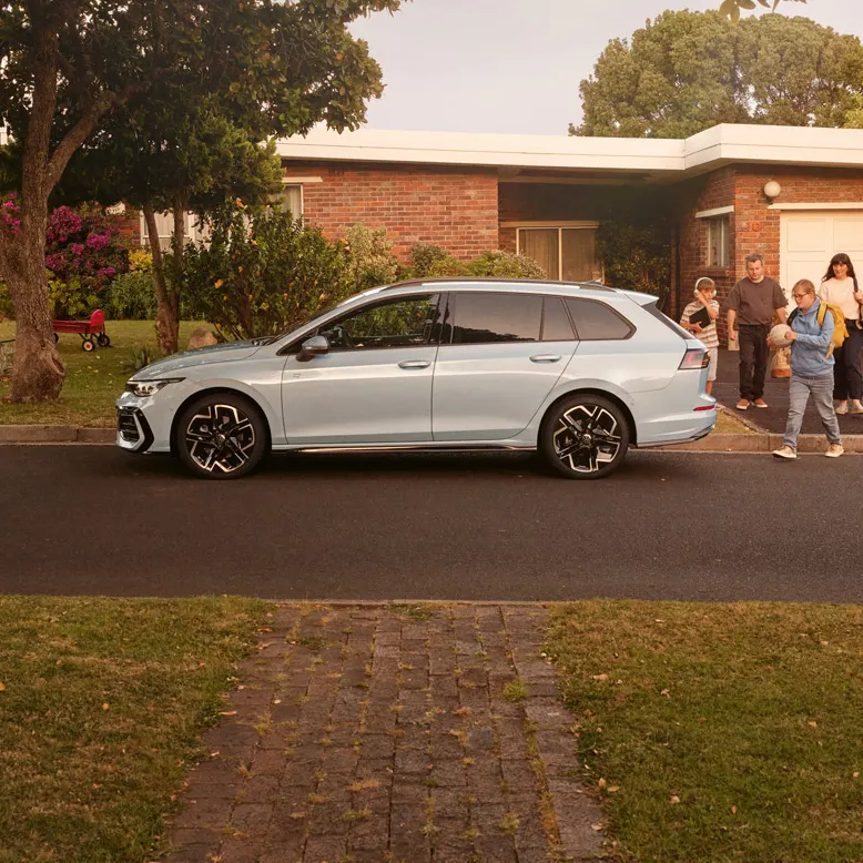 Ein weißer VW Golf Variant parkt vor einem Bungalow in einer Wohnstraße mit Bäumen. 