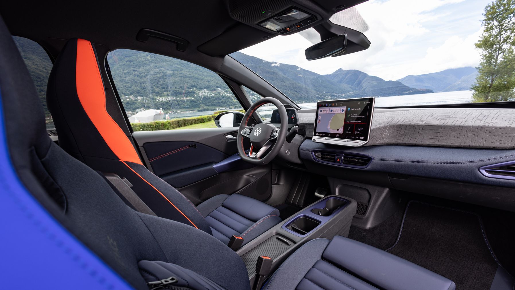 Das Bild zeigt den Innenraum eines modernen Autos mit zwei Frontsitzen, die in Dunkelblau und Orange gehalten sind. Die Mittelkonsole hat ein großes Display, das Navigations- und Infotainmentinformationen anzeigt. Im Hintergrund sind Berge und ein See zu sehen.