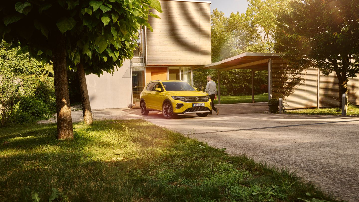 Ein gelber VW T-Cross steht in der Auffahrt eines modernen Wohnhauses mit großen Bäumen.
