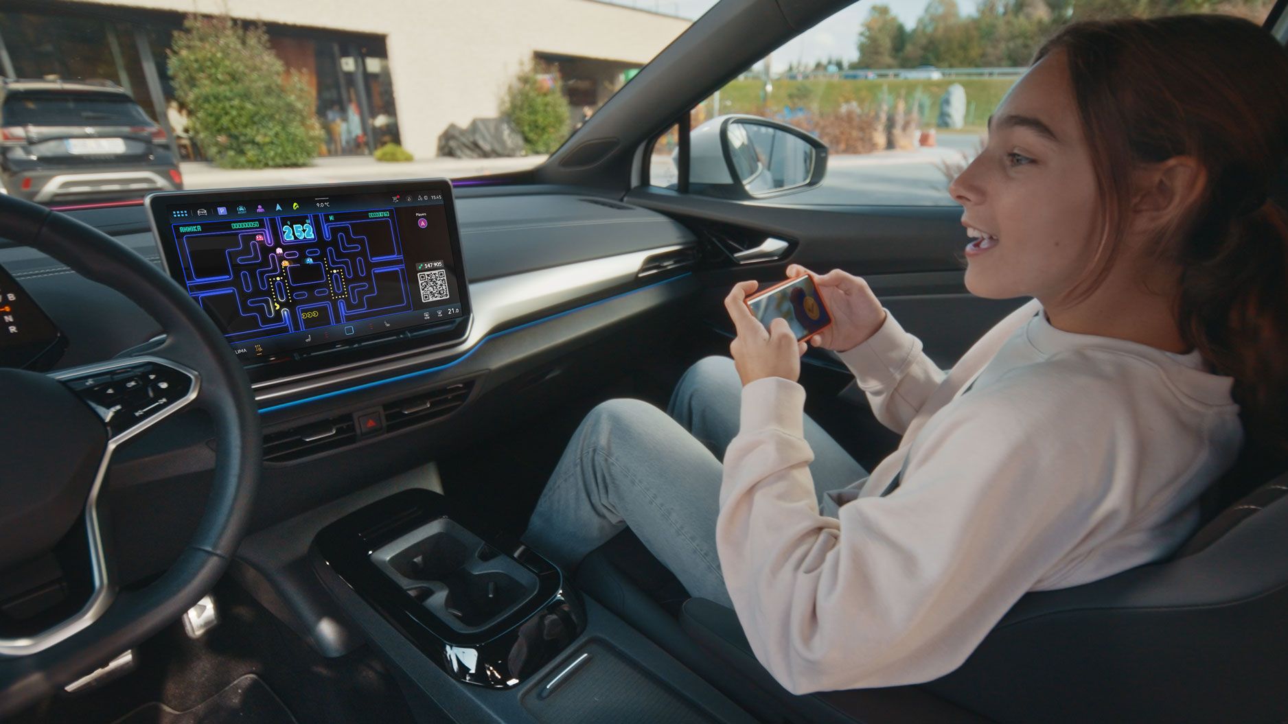 Eine Person sitzt entspannt im Fahrersitz eines Autos und spielt ein Videospiel auf ihrem Smartphone. Das Auto hat ein großes eingebautes Display auf dem Armaturenbrett, das ebenfalls ein Videospiel zeigt. Im Hintergrund ist die Außenseite eines Gebäudes zu sehen.