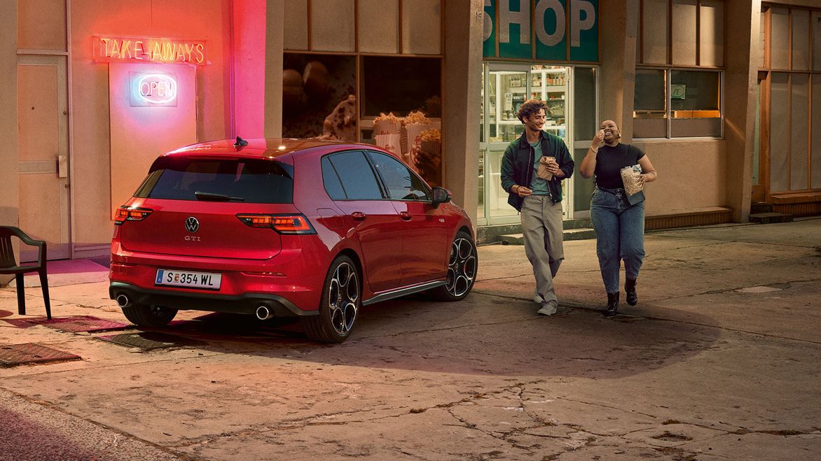  Heckansicht eines roten VW Golf GTI in abendlicher Stimmung, zwei Personen gehen neben ihm her