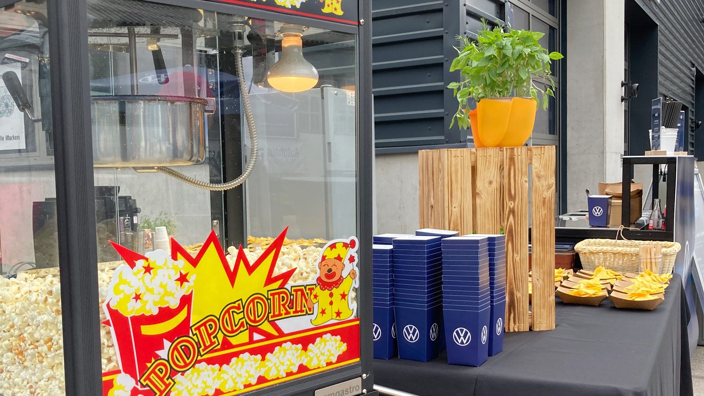 Eine Popcornmaschine mit frischem Popcorn steht auf einem Tisch. Daneben sind stapelweise blaue Pappbecher mit einem Volkswagen-Logo sowie ein Tisch mit Snacks und einer Topfpflanze. Im Hintergrund sind industrielle Gebäude zu sehen.