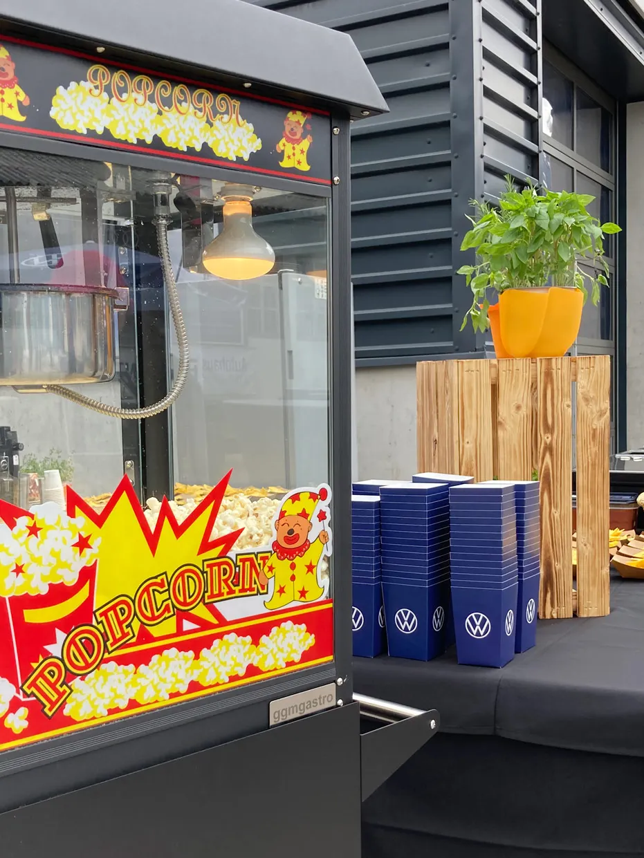 Eine Popcornmaschine mit frischem Popcorn steht auf einem Tisch. Daneben sind stapelweise blaue Pappbecher mit einem Volkswagen-Logo sowie ein Tisch mit Snacks und einer Topfpflanze. Im Hintergrund sind industrielle Gebäude zu sehen.