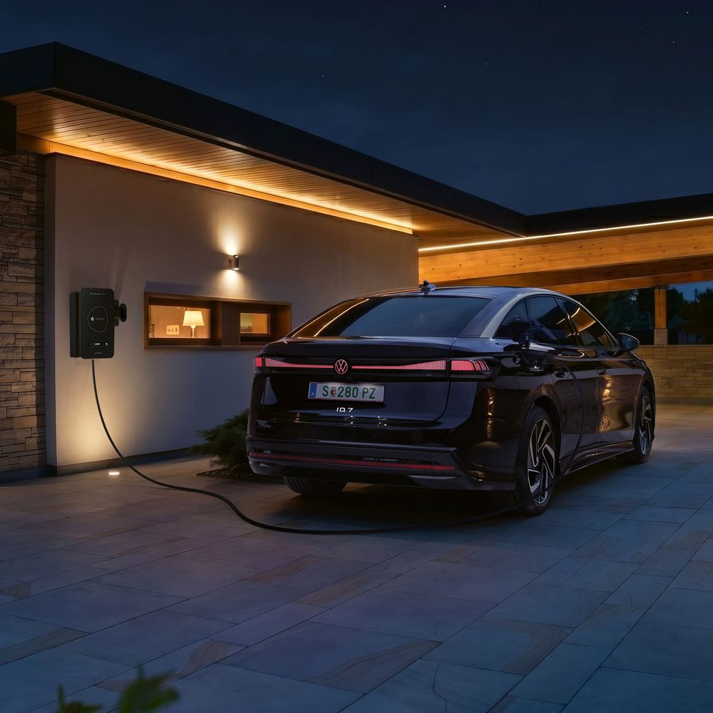 Ein schwarzes Elektroauto steht nachts in der Einfahrt eines modernen Hauses und ist an einer Wallbox zum Laden angeschlossen. Warmes, indirektes Licht an Fassade und Carport beleuchtet die Szene stimmungsvoll. Im Hintergrund sind Berge oder Hügel im Dämmerlicht zu erkennen.