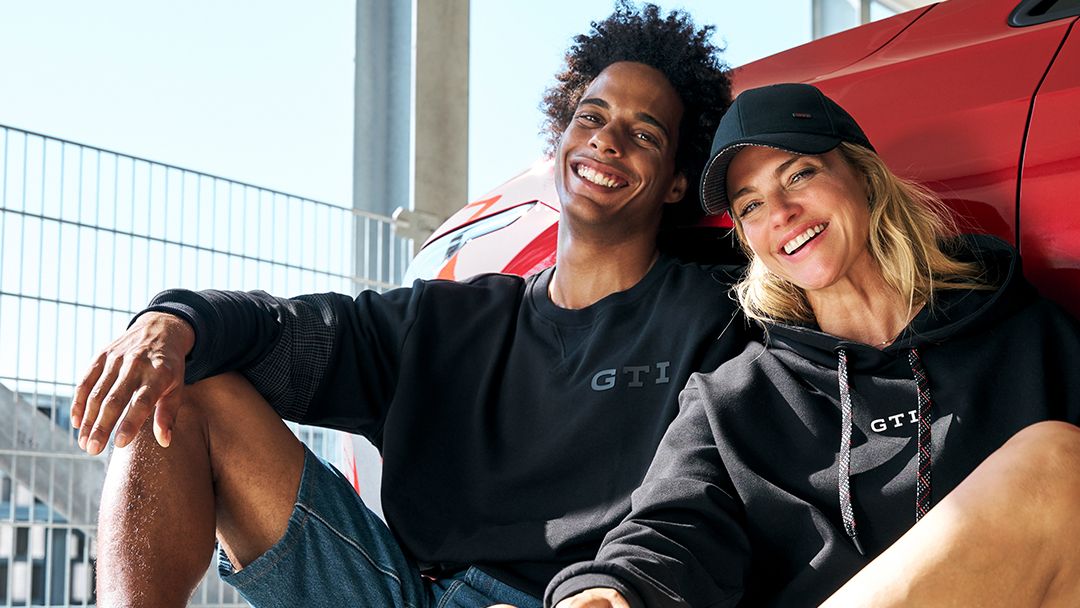 Zwei junge Menschen sitzen entspannt nebeneinander auf dem Boden vor einem roten Volkswagen und lächeln in die Kamera. Beide tragen lässige GTI-Merchandise-Kleidung mit Hoodies oder Sweatshirts, Shorts und Sneakern.