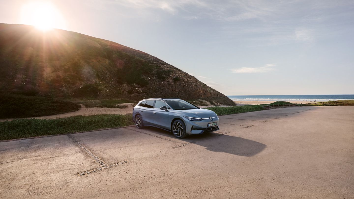Ein blauer VW ID.7 Tourer steht auf einem Parkplatz am Berg, im Hintergrund ein Strand und das Meer.