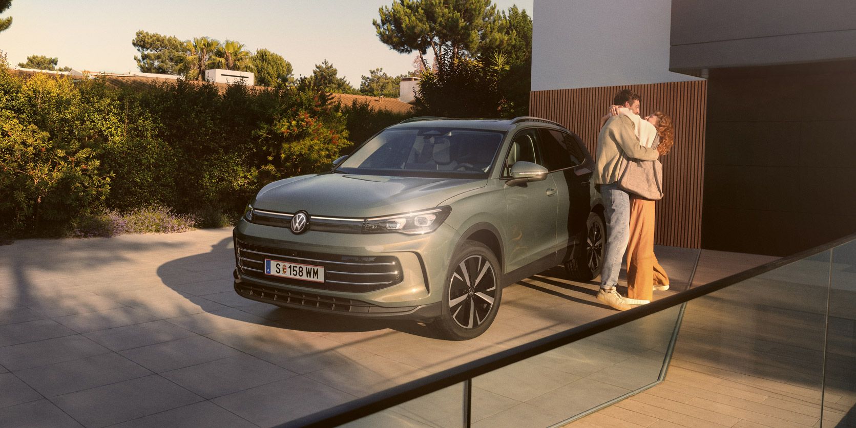 Eine Frau und ein Mann umarmen sich, neben ihnen steht ein VW Tiguan Elegance in der Farbe Cipressino-Grün Metallic