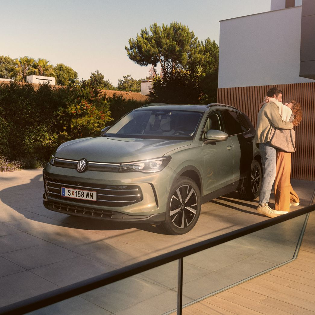 Eine Frau und ein Mann umarmen sich, neben ihnen steht ein VW Tiguan Elegance in der Farbe Cipressino-Grün Metallic