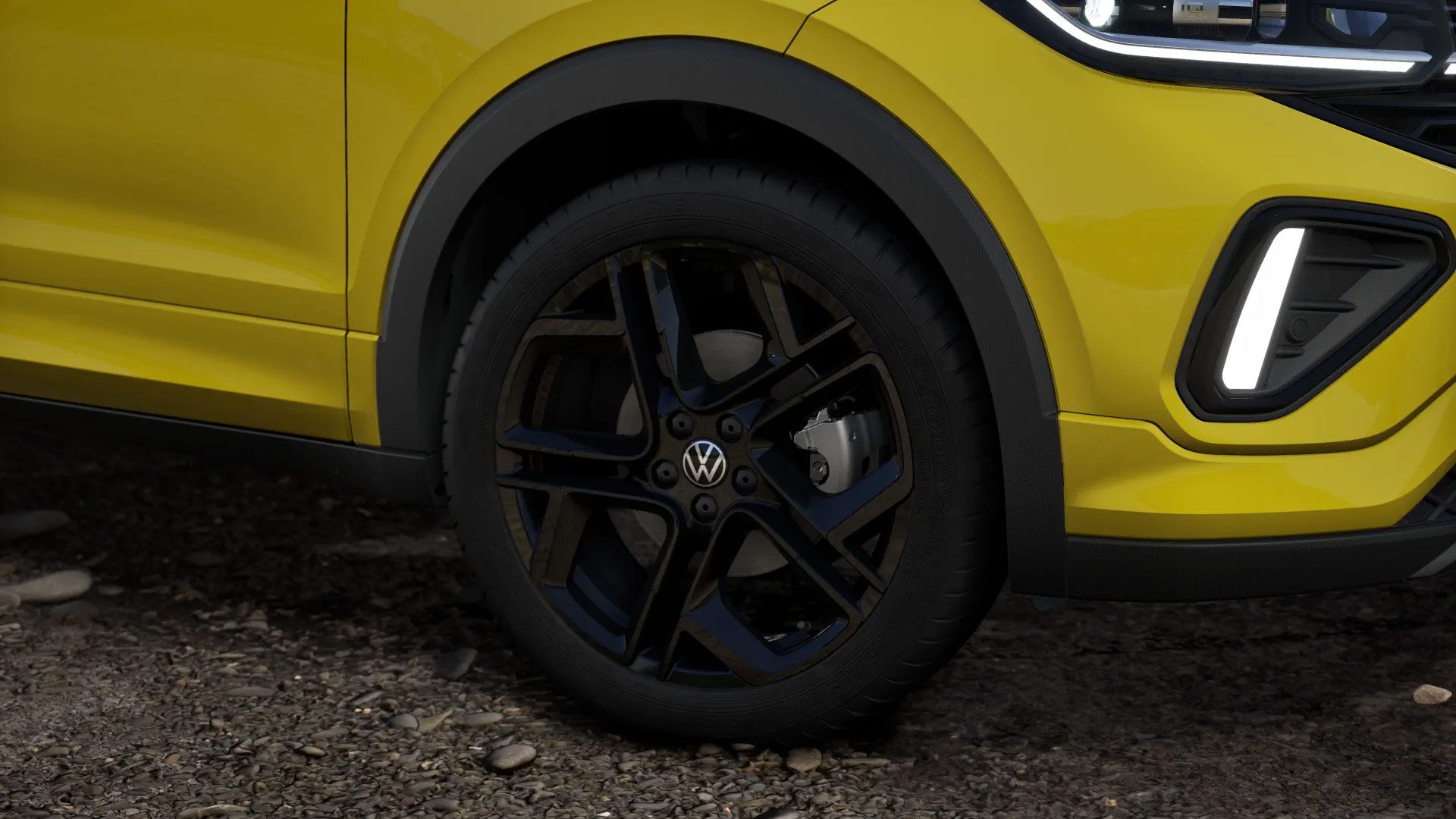 Nahaufnahme eines gelben Volkswagen T-Cross zeigt das vordere Rad und einen Teil der Karosserie. Die schwarzen Leichtmetallfelgen heben sich deutlich ab. Der Hintergrund besteht aus einem unbefestigten, kiesigen Boden.